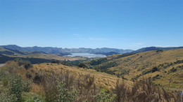 Wide scenic view of rolling golden hills and scrubland leading down to a blue lake in the distance, with layered mountain ranges on the horizon under a clear blue sky.