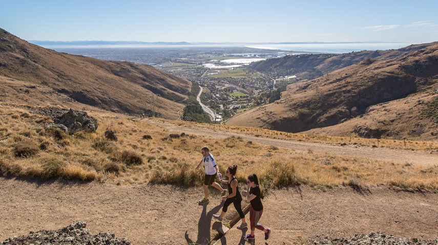 Three people walking along a dry hillside trail overlooking a valley town and coastline.