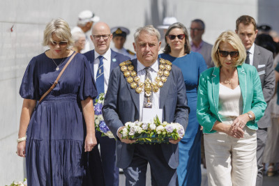 A group of people walk together during a memorial service, with the central figure, the mayor, carrying a floral wreath, while others walk alongside.
