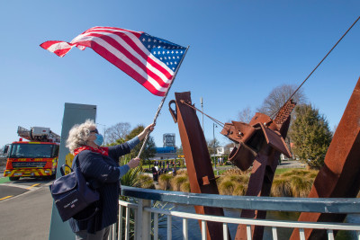 A person waves a United States flag beside a metal sculpture near a roadside, with emergency vehicles and buildings visible in the background.