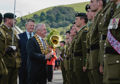 A mayor wearing a ceremonial gold chain shakes hands with uniformed soldiers standing in formation during an outdoor military ceremony, with a brass band and green hills in the background.