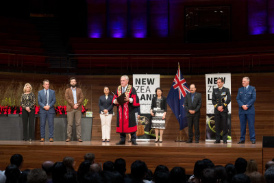 A group of people stand on a stage during a formal ceremony, with the mayor speaking in front of the New Zealand flag, and floral arrangements, while an audience sits below.