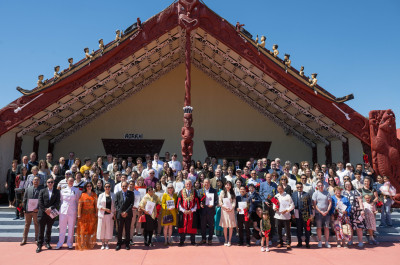 A large group of people pose for a photo outside a marae with carved details and red architectural features.
