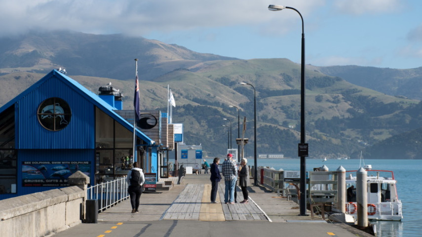 Akaroa's wooden wharf beside blue waterfront building with people walking and standing, boats docked nearby, and green hills in the background.