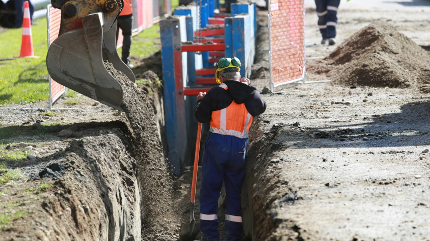 Construction worker in a trench as an excavator pours soil beside roadside barriers and cones.